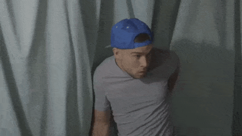 a man wearing a blue hat and a grey shirt is standing in front of a curtain
