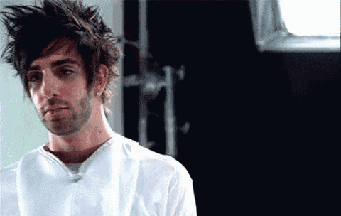 a man with messy hair and a beard is wearing a white shirt and looking at the camera .