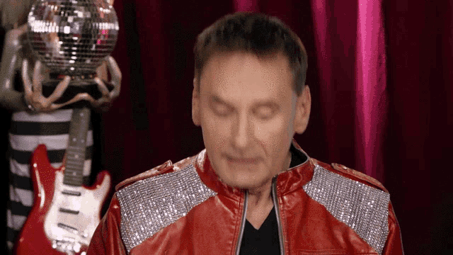 a man in a red jacket with rhinestones on the sleeves stands in front of a disco ball