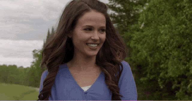 a woman in a blue shirt is smiling in front of a field of trees