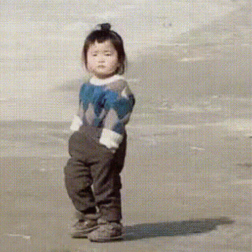 a little girl wearing a sweater and overalls is standing on a beach