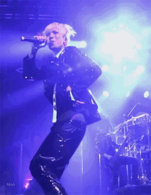 a man is singing into a microphone on a stage with a blue light behind him and the name mrick is on the bottom right