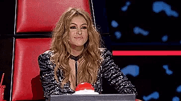 a woman in a sequined jacket is sitting in a red chair with a red button in front of her .