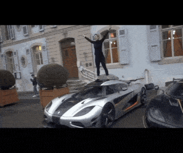 a woman stands on top of a sports car with her arms in the air