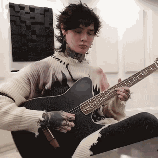 a man in a sweater is playing a guitar with the letter t on the neck