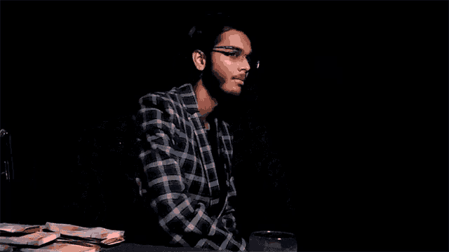 a young man wearing glasses and a plaid jacket is holding his hair .