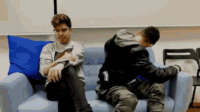 two men are sitting on a blue couch with one wearing a black jacket with the adidas logo on it