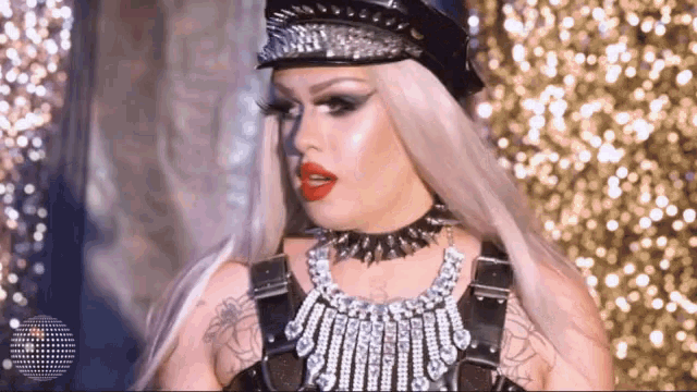 a drag queen wearing a hat and a necklace is standing on a stage .