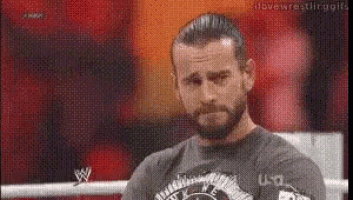 a man with a beard is sitting in a boxing ring wearing a gray shirt .