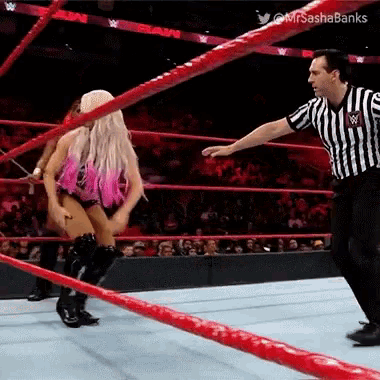 a woman in a pink outfit is dancing in a wrestling ring while a referee watches