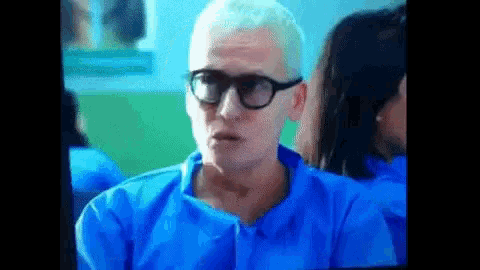 a man wearing glasses and a blue shirt is sitting in a room with other people .