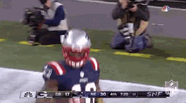 a football player in a patriots uniform is kneeling on the field while a photographer takes a picture of him .