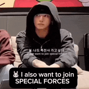 a man in a black hoodie is sitting on a couch with a sign that says i also want to join special forces