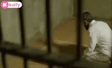 a man in a white shirt is sitting in a cell behind bars .