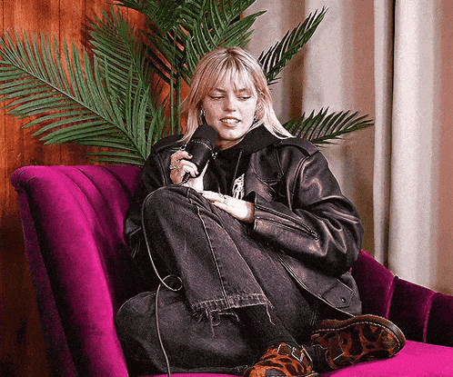 a woman in a leather jacket is sitting on a purple couch holding a microphone .