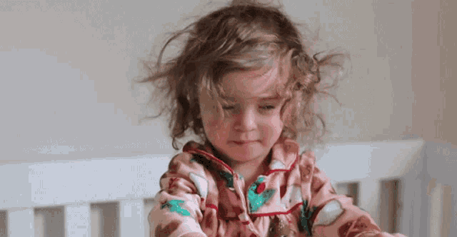 a little girl with curly hair is sitting in a crib and looking at the camera .