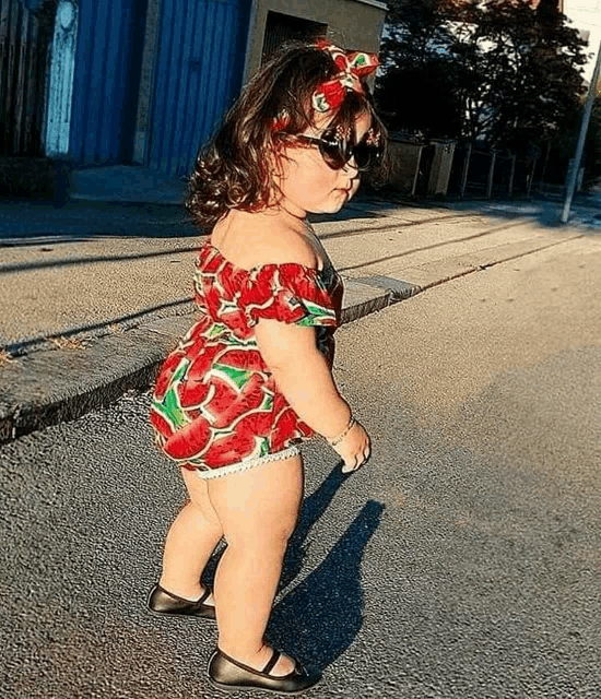 a little girl wearing a watermelon outfit and sunglasses is standing on the side of the road .