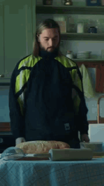 a man with long hair and a beard is standing in a kitchen next to a table with bread on it .