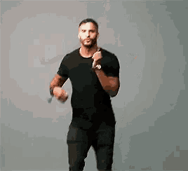 a man in a black shirt and black jeans is dancing in front of a gray background .