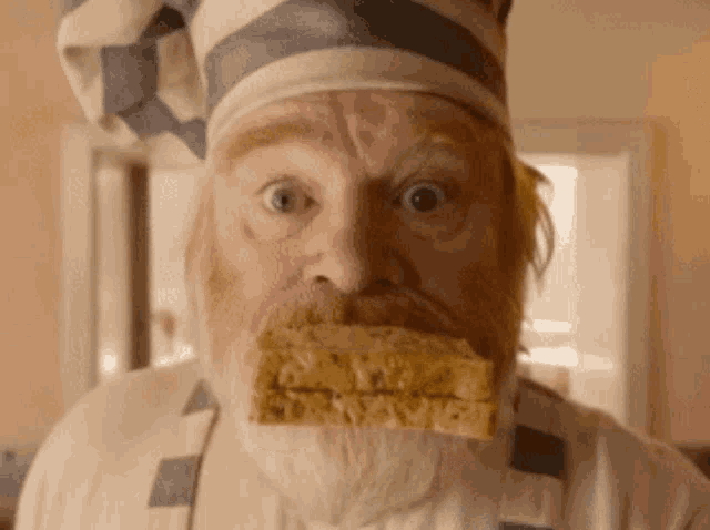 a man with a beard wearing a chef 's hat and apron is holding a piece of food in his mouth