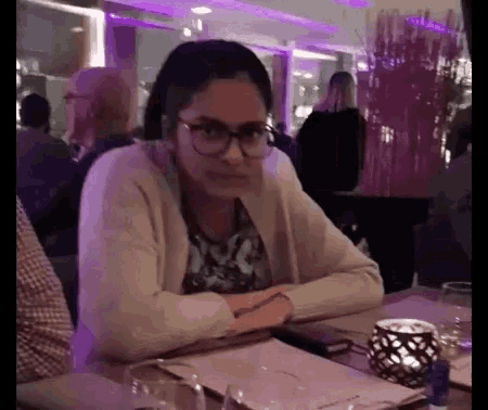a woman wearing glasses is sitting at a table in a restaurant with her hands folded .