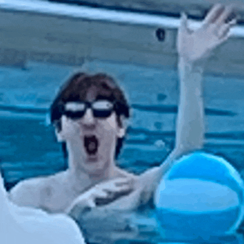 a man wearing sunglasses is swimming in a pool while holding a blue beach ball .