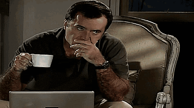 a man sitting in a chair holding a cup of coffee and looking at a laptop