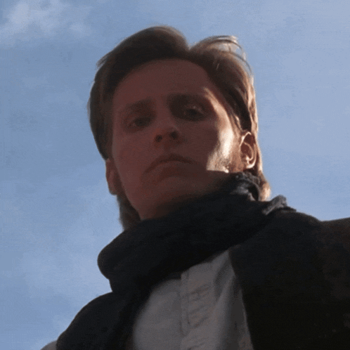a man wearing a scarf and a white shirt looks down at the camera