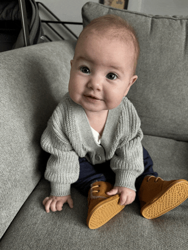 a baby wearing a grey cardigan and brown shoes sits on a grey couch