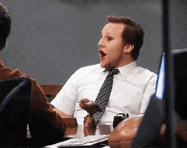 a man in a white shirt and tie is sitting at a desk with his mouth open .