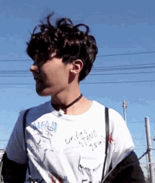 a young man wearing a white t-shirt and a choker is standing in front of a blue sky .
