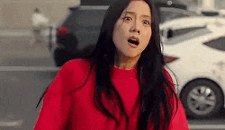 a woman in a red sweater is standing in front of a white car and making a surprised face .