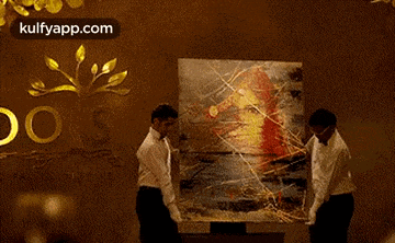two men are holding a large painting in front of a wall that says ' kulfyapp.com ' on it .