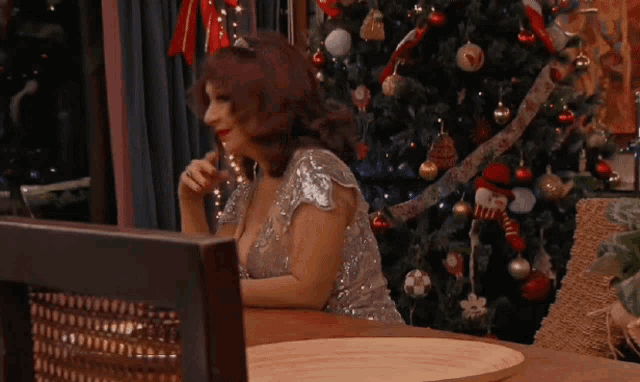 a woman in a sequined dress is sitting at a table in front of a christmas tree
