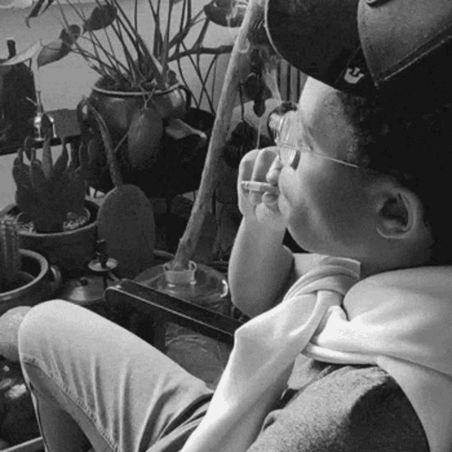 a black and white photo of a person wearing a hat and glasses smoking a cigarette .