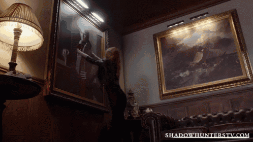 a woman hangs a painting in a room with shadowhunterstv.com on the bottom