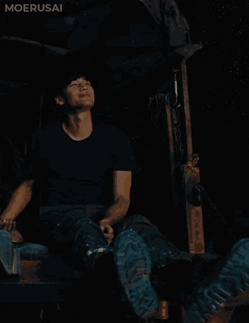 a man in a black shirt is sitting in a dark room with the word moerusai on the bottom right