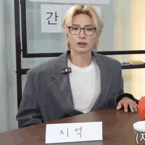 a man wearing glasses sits at a table with a name tag that says " 지석 " on it