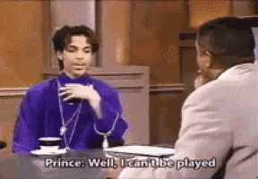 prince is sitting at a table talking to a man in a suit