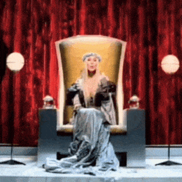 a woman in a long dress is sitting on a throne with a red curtain behind her .