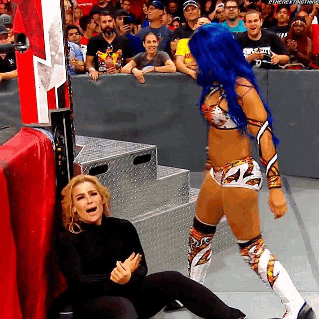 a wrestler with blue hair is standing next to another wrestler who is laying on the ground