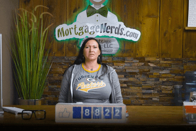 a woman wearing a green bay packers shirt sits at a desk in front of a sign that says mortgage nerds.com