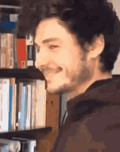 a man with a beard is smiling in front of a bookshelf with books on it .