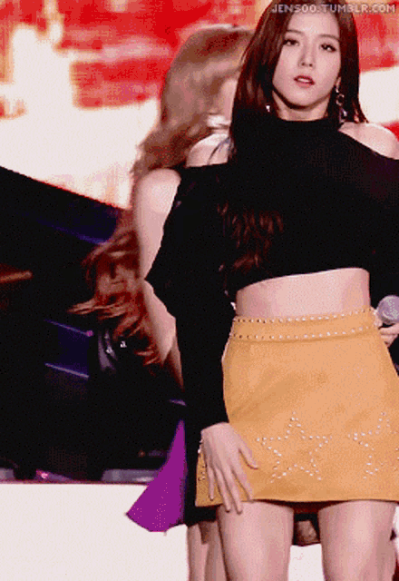 a woman in a black crop top and a yellow skirt is holding a microphone while another woman stands behind her