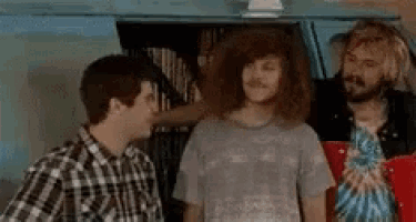 three men are standing next to each other in a room . one of the men has long curly hair .