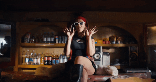 a woman wearing sunglasses is sitting on a table in front of a shelf full of soda bottles