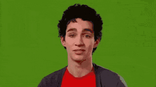 a young man with curly hair and a red shirt is making a funny face against a green screen .
