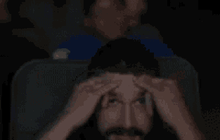 a man with a beard is sitting in a theater watching a movie and touching his forehead .