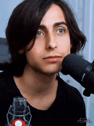 a young man with long black hair is standing in front of a shure microphone .