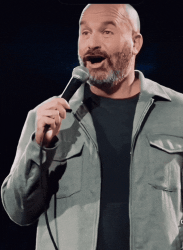 a man with a beard is singing into a microphone while wearing a grey jacket
